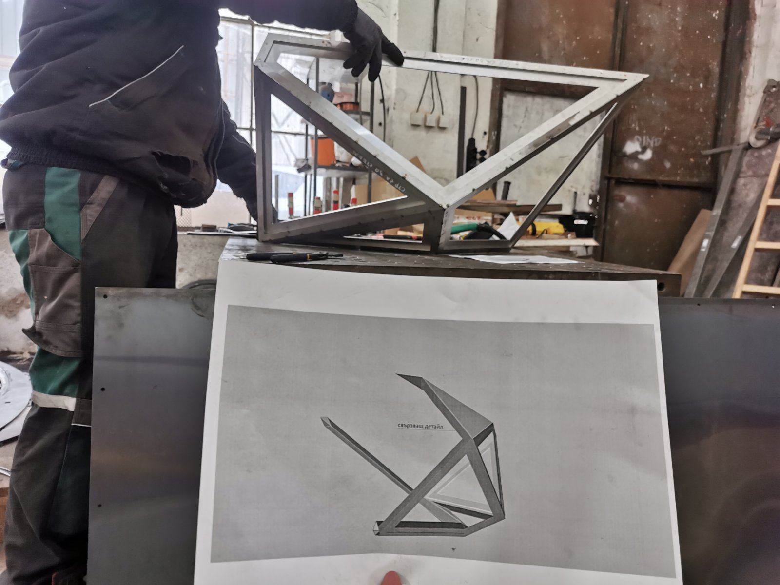 A welded frame held above its printed 3D drawing