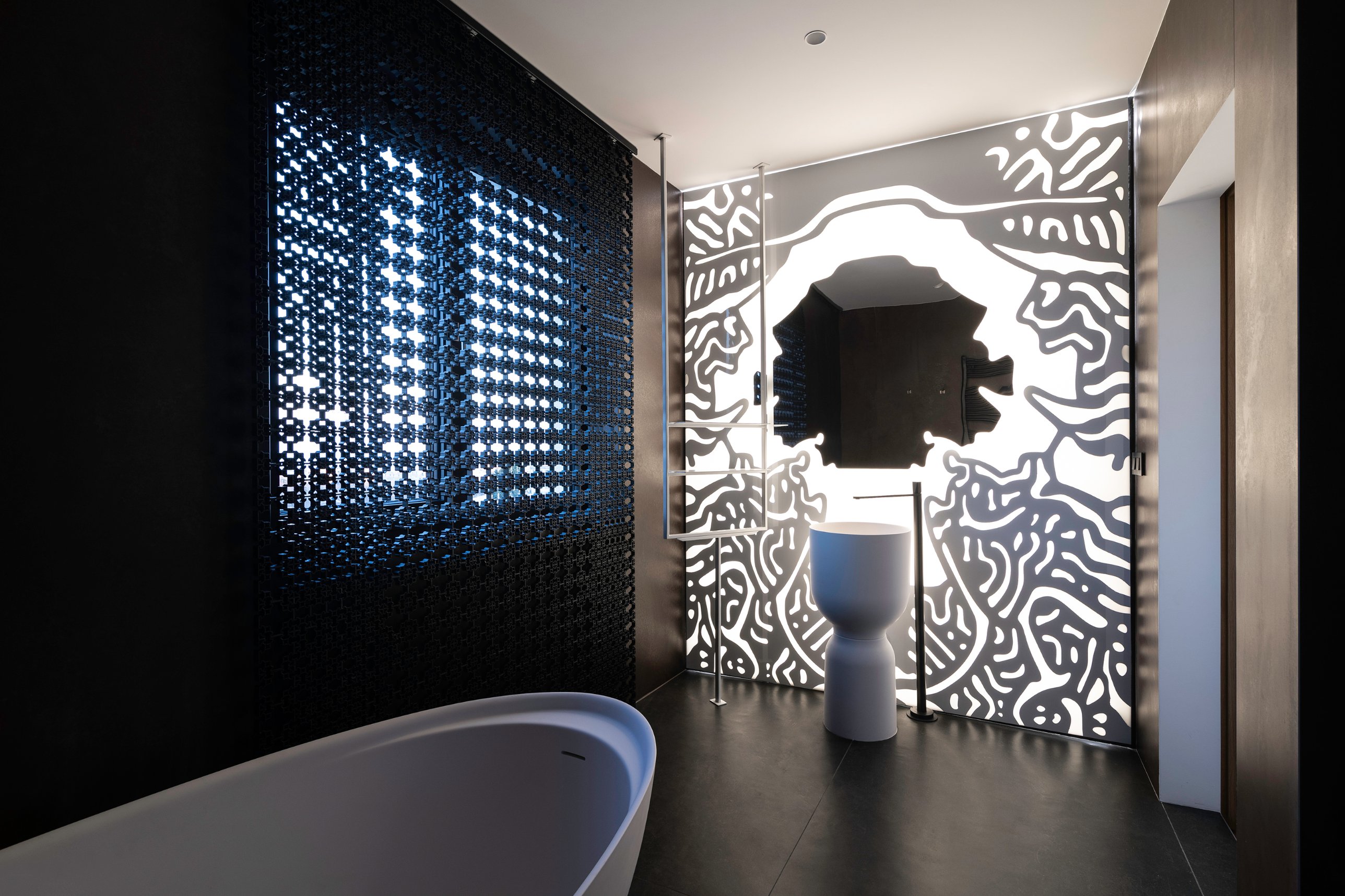 A freestanding white basin and bath before an illuminated abstract mirror wall and a perforated sliding panel