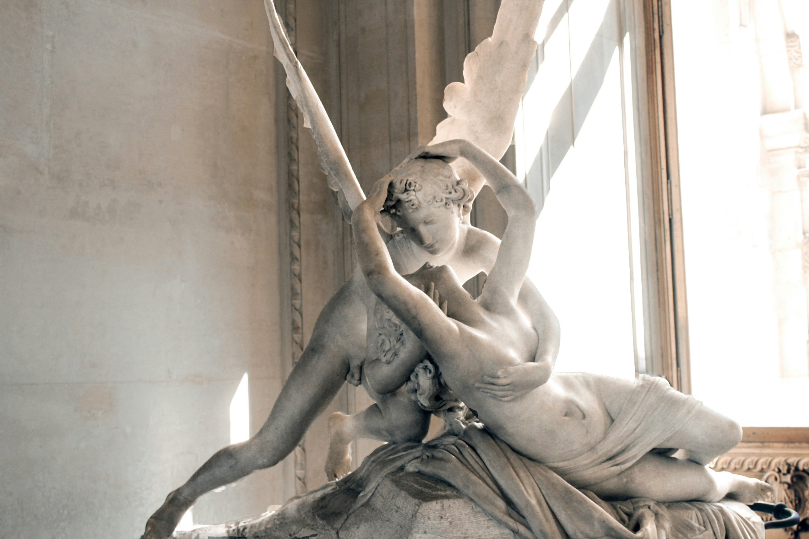 A white marble sculpture of a winged figure embracing a reclining figure, lit by a tall window